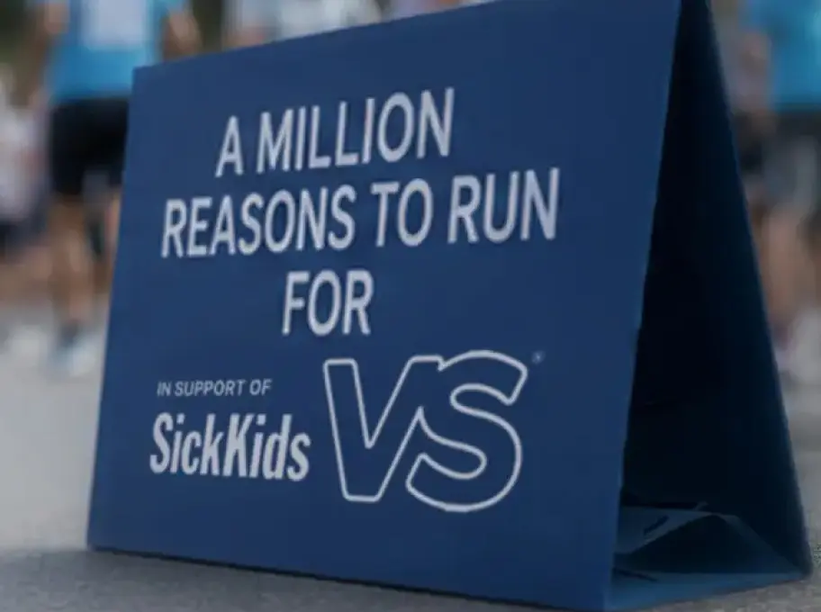 Sign promoting the SickKids Million Reasons to Run fundraising event with participants gathered in the background supporting community fundraising efforts.